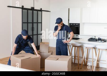 Nachdenklicher Arbeiter, der mit der Hand auf der Hüfte neben Kollegen und Kartonschachteln in der Küche steht Stockfoto