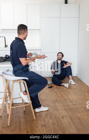 Fröhlicher Handwerker, der mit einem Kollegen spricht, während er auf dem Boden in der Küche sitzt Stockfoto