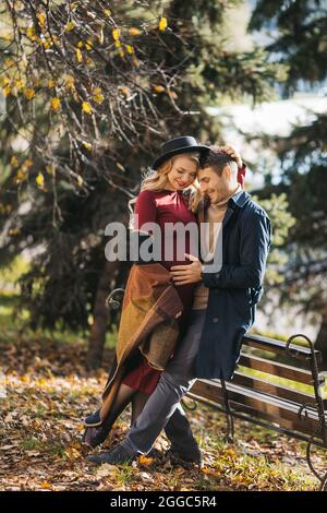 Junge kaukasische glückliches Paar posiert draußen am Herbsttag. Eine junge Schwangere umarmt ihren Mann und posiert im herbstlichen Stadtpark Stockfoto