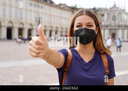 COVID-19 optimistische Reisende Frau trägt schwarze Schutzmaske KN95 FFP2 zeigt Daumen nach oben in der Stadt Platz Stockfoto