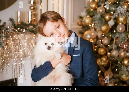 Der kaukasische Junge umarmt seinen geliebten Hund auf dem Hintergrund eines weihnachtsbaums zu Hause. Glückliches Kind an den weihnachtsferien. Haustiere im Urlaubsdekor Stockfoto