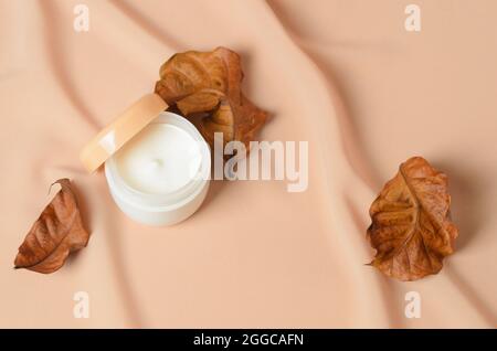 Ein Glas Creme für die trockene Hautpflege mit getrockneten Herbstblättern auf einem welligen beigen Hintergrund aus Chiffon. Selektiver Fokus. Stockfoto