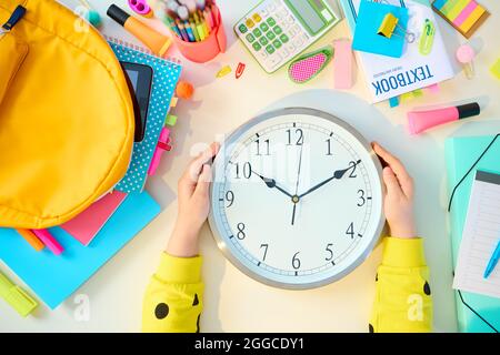 Zurück zur Schule. Obere Ansicht des modernen Mädchens mit Arbeitsbüchern, Schreibwaren, Lehrbuch, Uhr und Rucksack am weißen Tisch zu Hause. Stockfoto