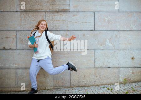 Happy moderne Schulmädchen in weißen Sweatshirt mit Arbeitsbuch und Rucksack springen und gegen die Wand im Freien in der Stadt. Stockfoto