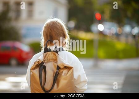 Von hinten gesehen trendiges Schulmädchen im weißen Sweatshirt mit Rucksack, der über den Crosswalk geht und in der Stadt draußen zur Schule geht. Stockfoto