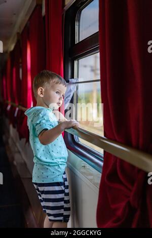 Nachdenklicher kleiner Junge, der durch das Fenster mit dem Zug auf einen bequemen Eisenbahnwagen schaut Stockfoto