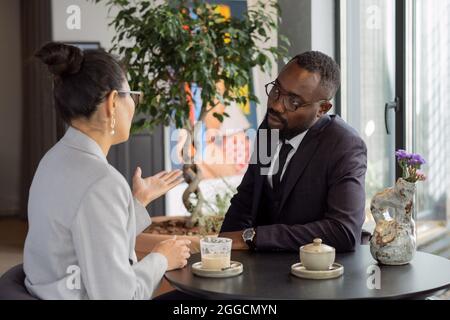 Zwei junge interkulturelle Makler bei einem Drink und einem Gespräch über Arbeitsmomente bei einem Treffen im Café Stockfoto