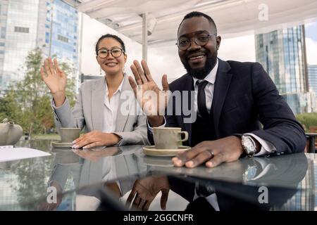 Asiatische und afrikanische Büroangestellte winken ihren Kollegen während eines Online-Meetings vor dem Laptop im Café im Freien die Hände Stockfoto