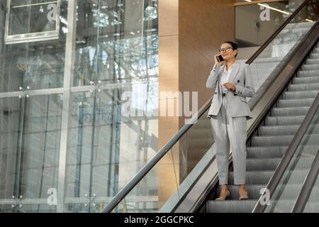 Glückliche Geschäftsfrau in grauem Anzug, die auf dem Smartphone spricht, während sie sich auf der Rolltreppe nach unten bewegt Stockfoto