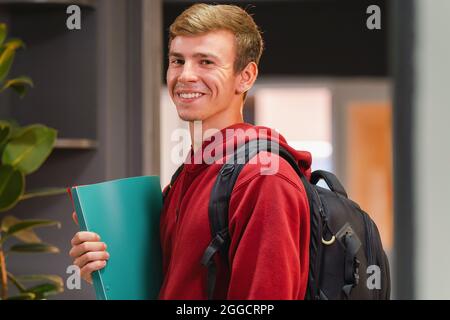 Seitenansicht Foto eines glücklichen Studenten in der Universität in einem roten Hoodie mit Rucksack auf der Rückseite, der die Farbmappe in der Hand hält. Stockfoto