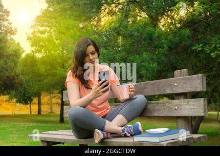Schöne junge hispanische Frau, die auf einer Holzbank im Park sitzt, in Sportkleidung gekleidet, mit einer Tasse Kaffee in der Hand, um eine Botschaft auf ihren s zu lesen Stockfoto