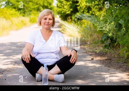 Nachdenkliche Frau, die sich auf der Straße ausruht, mit gekreuzten Beinen in Sportkleidung, die ihre Hände auf den Knien hält, befindet sich die Flasche Wasser in der Nähe der Frau. Stockfoto