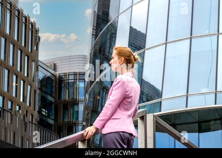 Die junge Blondine mit maßgeschneiderter Frisur in pinker Jacke steht auf einer Treppe und blickt auf ein modernes Bürogebäude aus Glas und Stahl. Profilansicht. Sel Stockfoto
