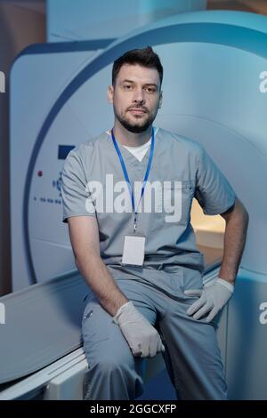 Selbstbewusster Arbeiter des medizinischen Labors, der vor der Kamera auf dem Tisch der CT-Geräte sitzt Stockfoto