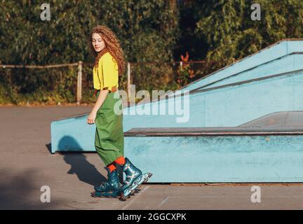 Junge rothaarige Frau in grünen und gelben Kleidern und orangefarbenen Strümpfen mit lockigen Frisuren auf Rollerblades im Skate Roller Park laufen Stockfoto