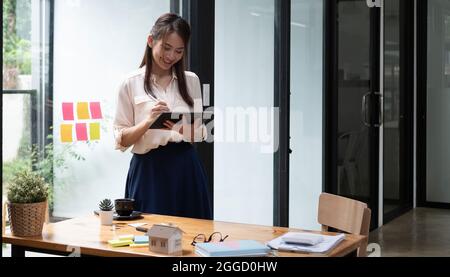 Glückliche Managerin hält Tablet und steht im modernen Büro. Asiatische Frau mit Stift mit Tablet. Business Financial Accounting Konzept Stockfoto