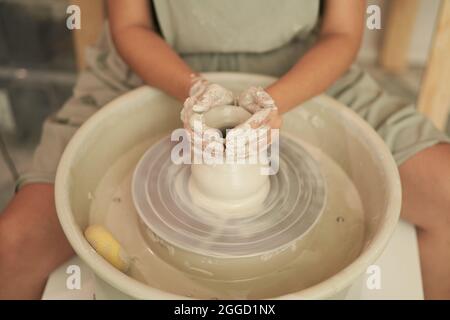 Anonyme Töpferin mit hohem Erntewinkel und schmutzigen Händen, die Tontopf auf rotierendem Rad modelliert, während sie in einem Kunsthandwerkerstudio arbeitet Stockfoto