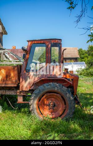 Ein alter rostiger Traktor steht in der Nähe der Scheune. Auf dem grünen Gras steht ein roter alter Traktor für den Haushalt und die Ernte. Stockfoto