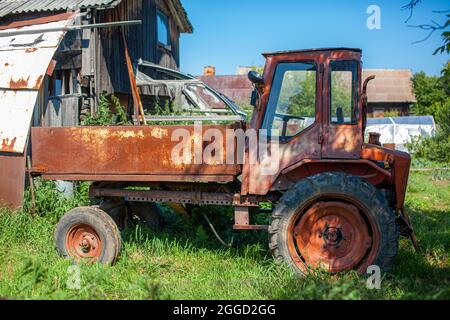 Ein alter rostiger Traktor steht in der Nähe der Scheune. Auf dem grünen Gras steht ein roter alter Traktor für den Haushalt und die Ernte. Stockfoto