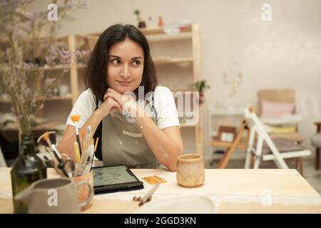 Lächelnde, verträumte junge Künstlerin in Schürze, die mit Tablet und Tongeschirr am Tisch sitzt und im Kreativstudio über neues Design für Geschirr nachdenkt Stockfoto