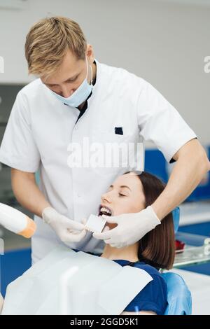 Nahaufnahme Porträt der schönen jungen Dame auf dem Zahnstuhl sitzen, während Stomatologe Hände in sterilen Handschuhen halten Zahnproben. Sie lächelt Stockfoto