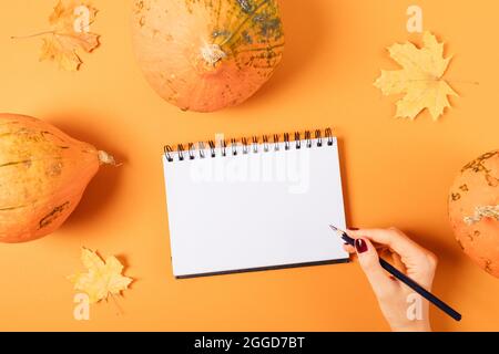 Weibliche Hand schreibt in Notizbuch mit leerem weißen Blatt zwischen Kürbissen und Herbstblättern auf orangefarbenem Hintergrund, flach liegend. Stockfoto