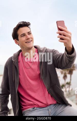 Vertikales Foto eines glücklichen jungen Mannes, der ein Selfie-Foto mit seinem Smartphone vor der Stadt macht, Konzept der Technologie und des urbanen Lebensstils, Kopie s Stockfoto