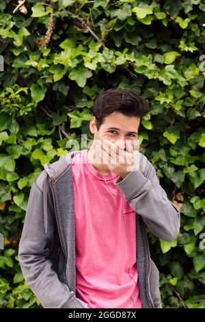 Vertikales Foto eines netten jungen Mannes, der lachend seinen Mund mit einer Hand auf einem grünen Pflanzenhintergrund bedeckt, Konzept von lustigen Ausdrücken und Emotionen, Co Stockfoto