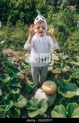 Niedlicher kleiner Junge im Halloween Geisterkostüm, der Spaß im Garten in der Nähe von Kürbissen hat. Stockfoto