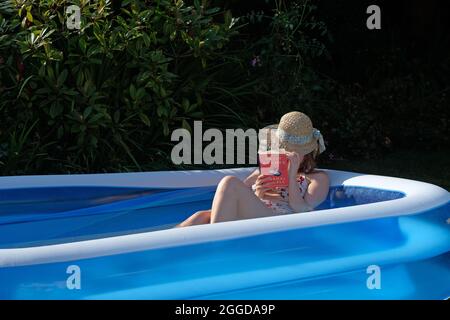 Eine junge Frau, die einen Sonnenhut trägt, liest ein Buch in einem Plastikplanschbecken in ihrem Garten Stockfoto