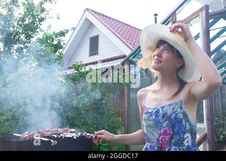 Ein Mädchen in einem Strohhut bereitet an einem hellen Sommertag vor grünem Laub aromatisches frisches Fleisch auf dem Grill zu. Nahaufnahme Stockfoto