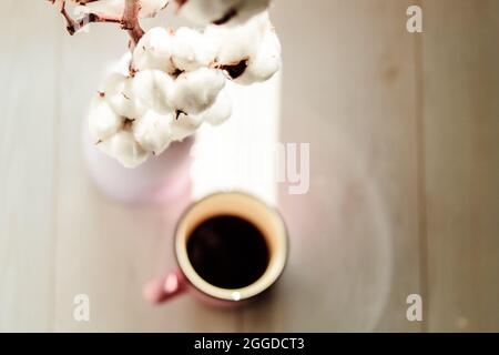 Eine Tasse schwarzen Kaffee in einer Keramikschale, ein Becher in Unschärfe, ein Zweig aus weißer Baumwollblume in einer Vase im Fokus auf einem weißen Parkettboden. Gemütliches h Stockfoto