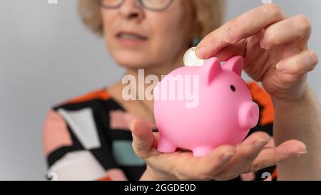 Eine ältere Frau mit Brille legt eine Münze in ein Sparschwein auf weißem Hintergrund Stockfoto