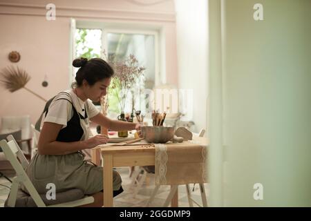 Seitenansicht einer kreativen jungen Frau im Vorfeld, die mit verschiedenen Werkzeugen am Tisch arbeitete, während sie im Studio Tonteller schuf Stockfoto