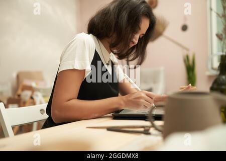 Seitenansicht der jungen weiblichen Keramikmeisterin mit Tablet und Stylus-Stift, während sie Design für Geschirr während des Arbeitsprozesses in der Werkstatt skizzierte Stockfoto