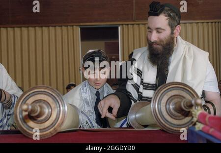 Ein Barmitzvah-Junge erhält seine erste Aliyah (aufgerufen, um aus der Schriftrolle der Tora zu lesen) während eines Dienstes an Wochentagen, wenn Tephillim (Lederkisten und Riemen auf dem Kopf getragen und ein Arm, der hebräische Schrift aus der Tora enthält) mit einem Tallit (einem Schal-Kleidungsstück) getragen wird. Von der männlichen Gemeinde. Mit dem Barmitzvah-Jungen ist der Rabbi, der ihn durch die Lesung führt. Diese Veranstaltung fand in einer regionalen Synagoge in den West Midlands, Großbritannien, statt Stockfoto