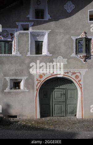 Weiße Sgraffitodekoration mit orangefarbenen Details ergänzt die grünen Holzvertäfelungen der Stalltür zu restauriertem Haus in Scuol, Graubünden oder Bündner Kanton, Schweiz. Das Haus aus dem Jahr 1726 verfügt über ein doppeltes, ebenfalls grün lackiertes „Balcun Tort“- oder Erkerfenster (rechts). Balcun Tort ist ein romanischer Begriff und diese Fenster sind typisch für die romanisch sprechende Schweiz. Stockfoto