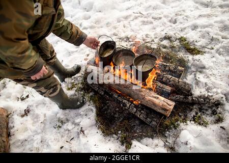 Drei Töpfe mit kochendem Wasser an einem Lagerfeuer steht an einem Wintertag ein Mann in warmen Kleidern neben ihm. Kochen auf der Wanderung. Stockfoto