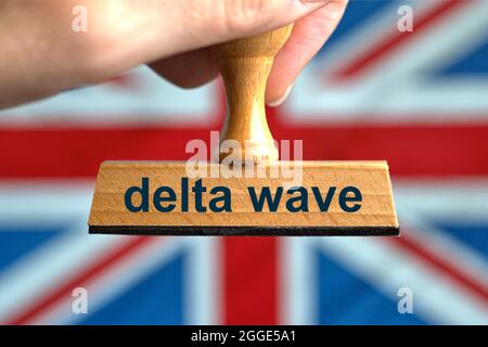 Deutschland. Juli 2021. Ein symbolischer Holzstempel mit der Aufschrift 'Delta Wave', der von einer Hand vor einer verschwommenen Flagge Großbritanniens (GB) gehalten wird, im Hintergrund der 'Union Jack'. Großbritannien hob am 19. Juli 2021 trotz hoher und steigender Inzidenzwerte alle Corona-Maßnahmen und -Beschränkungen für den „Freedom Day“ auf. Foto mit Komposition [M]. | Model Released Credit: dpa/Alamy Live News Stockfoto