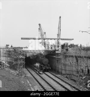 Ein Blick auf den Bau von Abschnitt A der Autobahn Birmingham nach Preston (M6), der zeigt, wie ein Team von Arbeitern einen der beiden 22-Tonnen-Stahlträger an der Eisenbahnbrücke Stafford/Wolverhampton (Brücke 302) mit einer Dampfmaschine, die unten auf der Eisenbahnlinie vorbeifährt, in Position senkt. Dieses Foto wurde im Mai 1961 in Laings monatlichem Newsletter „Team Spirit“ veröffentlicht. Die Arbeiten auf der Autobahn Birmingham-Preston (M6) zwischen den Kreuzungen J13 und J16 begannen im Juni 1960 und wurden von John Laing Construction Ltd. Durchgeführt.Abschnitt A war 51/4 Meilen lang zwischen Dunston und Whitgreav Stockfoto