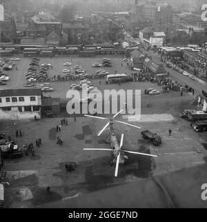 Blick vom Dach der Kathedrale von Coventry auf einen Hubschrauber der RAF Belvedere, der unten auf dem Asphalt geparkt ist, und zeigt eine Menschenmenge, die sich versammelt hat, um die Bushaltestelle Pool Meadow nördlich der Kathedrale zu beobachten. Das Foto wurde während der „Operation Rich man“ aufgenommen, einem gemeinsamen Projekt, an dem Mitarbeiter der Royal Air Force und Mitarbeiter von Laing beteiligt waren. Dazu wurde ein RAF Belvedere Hubschrauber hochgezogen, um den 80 Fuß großen Bronzeturm auf der neuen Kathedrale zu platzieren. Der Teil der Operation zur Senkung des 1/2 Tonnen schweren Kreuzes, das auf dem Turm sitzt, musste aufgrund windiger Bedingungen verschoben werden. Stockfoto