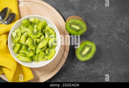 Kiwifruit frischer Sommersalat mit in Scheiben geschnittenen grünen Kiwi in einer weißen Schüssel auf einem Holzschneidebrett auf dunkelgrauer Tischansicht mit Kopierfläche. Stockfoto