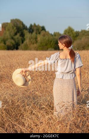 Ein Mädchen hält einen Hut in der Hand vor dem Hintergrund eines Weizenfeldes Stockfoto