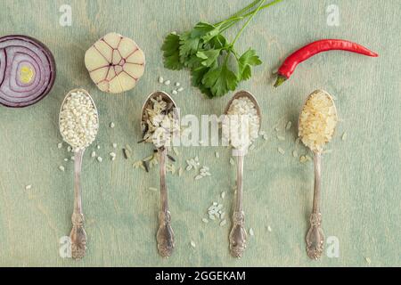 Flaches Lay verschiedener Reissorten in Löffeln mit Gewürzen. Wildreis, Sushi-Reis, geschredderter und gedämpfter Reis auf grünem Holzgrund mit Knoblauch, ch Stockfoto