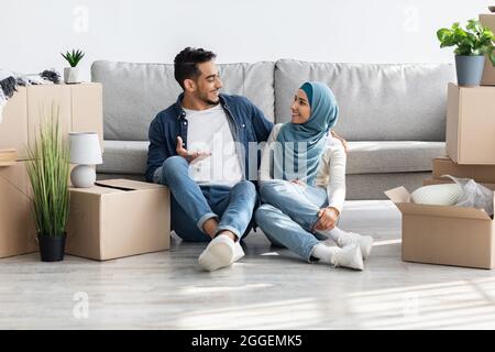 Aufgeregtes Paar aus dem Nahen Osten, das sich beim Auspacken nach dem Umzug ausruhte, glücklicher arabischer Kerl und eine Dame im Hijab, die auf dem Boden am Sofa zwischen Papierkisten saßen Stockfoto