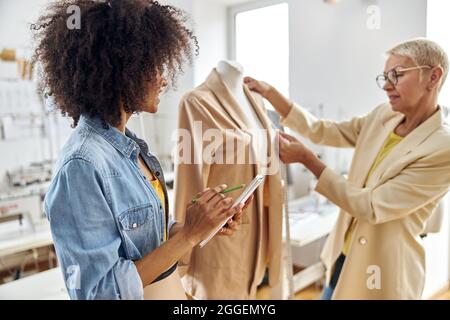 Afroamerikanische Frau zeichnet Skizzenbuch, während reife Schneiderin in der Werkstatt die Jacke misst Stockfoto