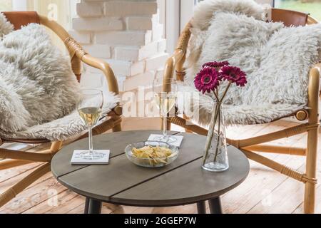 Gemütliche Szene am späten Nachmittag: Zwei Korbsessel mit Kissen und ein Couchtisch mit Weingläsern und ein Snack, zubereitet als Sundowner im Wintergarten Stockfoto