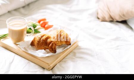 Croissant und Kaffee auf weißem Bett auf Korbtablett mit Blumen Stockfoto