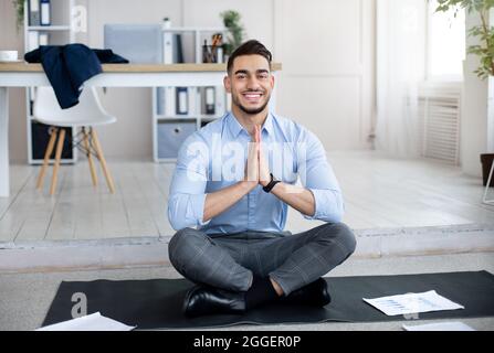 Stressmanagement am Arbeitsplatz. Friedlicher arabischer Mitarbeiter meditiert und macht im Büro eine namaste Geste auf der Yogamatte. Junger Mann aus dem Osten, der sich ruhig und balaneseim fühlt Stockfoto