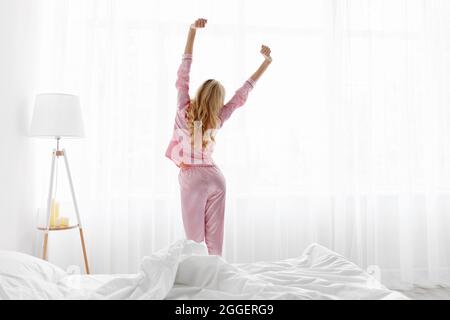 Frau wacht auf und steht vor dem Fenster und trifft den Sonnenaufgang. Glücklich schöne tausendjährige kaukasische Weibchen in rosa Pyjamas Stretching ihren Körper, in der Nähe weiß Stockfoto